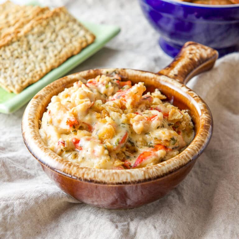 A brown crock filled with a dip with lobster poking out. There are crackers and chips in the background