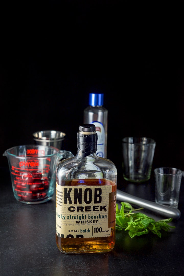 Bourbon, mint, cherries and simple syrup with a metal cup and measuring glasses in the background