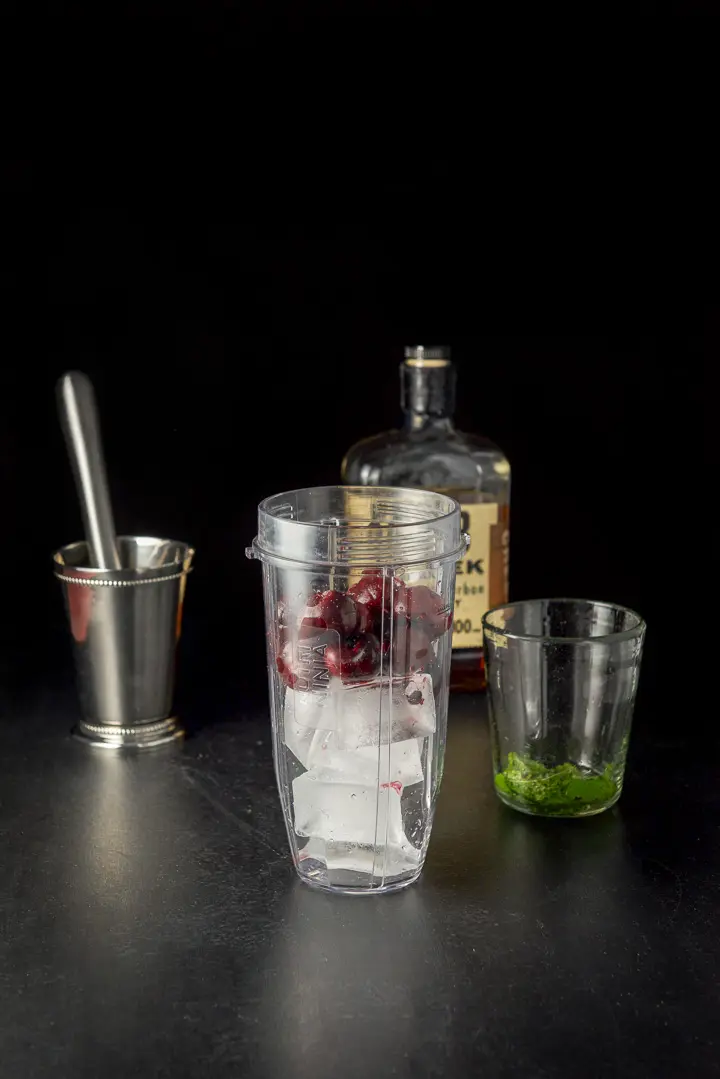 Ice cubes and pitted cherries in a blender container along with the metal cup, glass and bourbon in the background