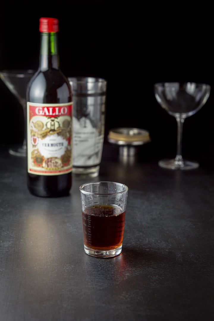 sweet vermouth poured out with the bottle, glasses and shaker in the background