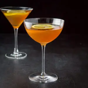 A bowl martini glass filled with the cocktail in front of the classic glass. There are lemon wheels in the drink as garnish - square