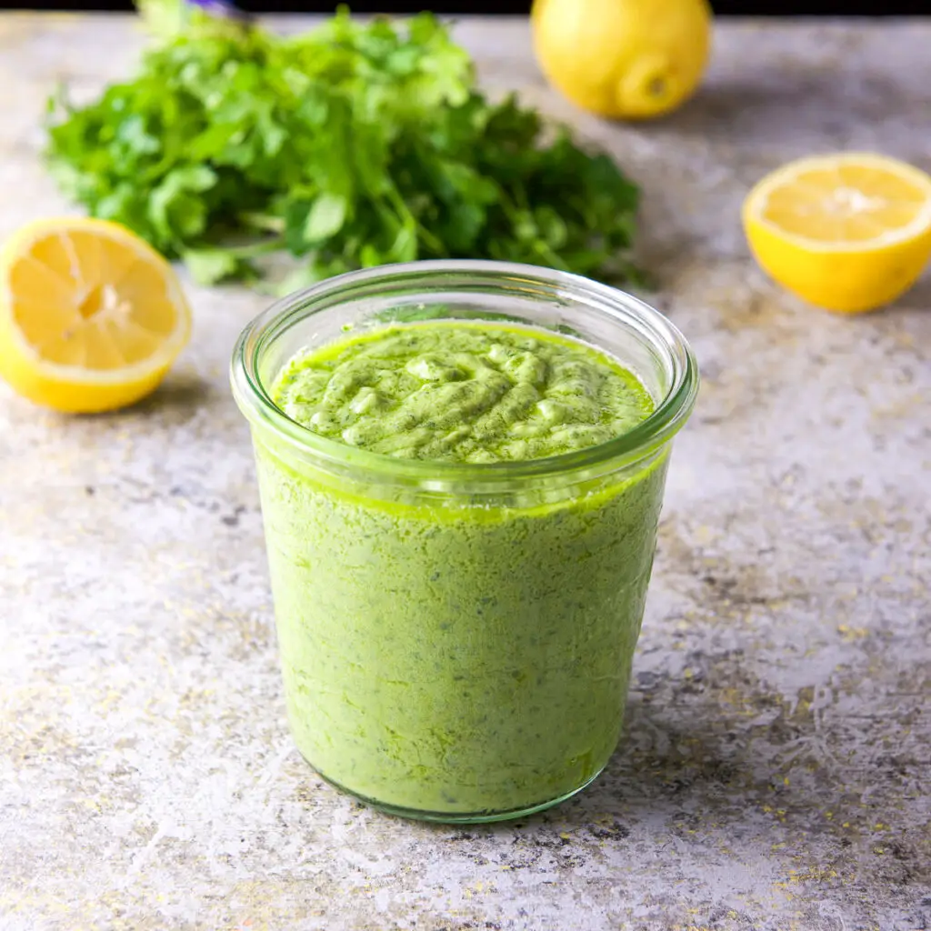 A jar with avo spread in it along with lemons and parsley in the background - square