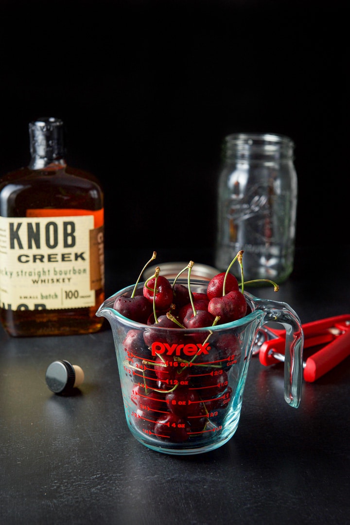 Cherries in a measuring cup, bourbon and a jar on a black table