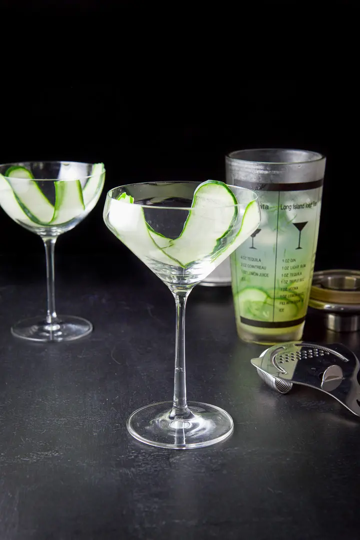 Thin cucumber slices draped in the glasses with the shaker filled with the cocktail in the background