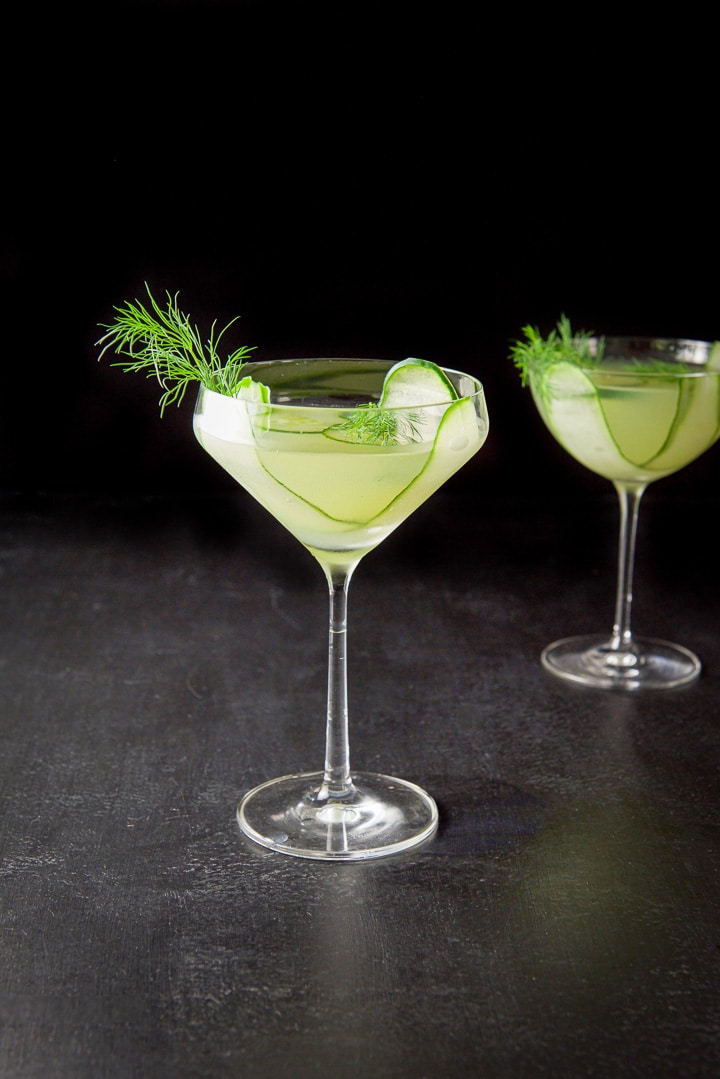 Curved glass with the cucumber martini in front of the bowl glass in the background. There is cucumber and dill as garnish