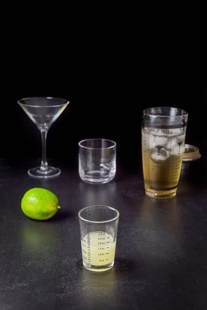 Lime juice poured out with a lime, shaker and glassware in the background