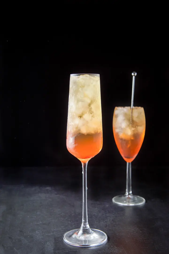 Tall champagne glass filled with the orange cocktail in front of the other glass with the stirrer