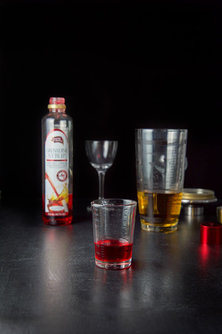 Grenadine measured out with the bottle, shaker and glasses in the background