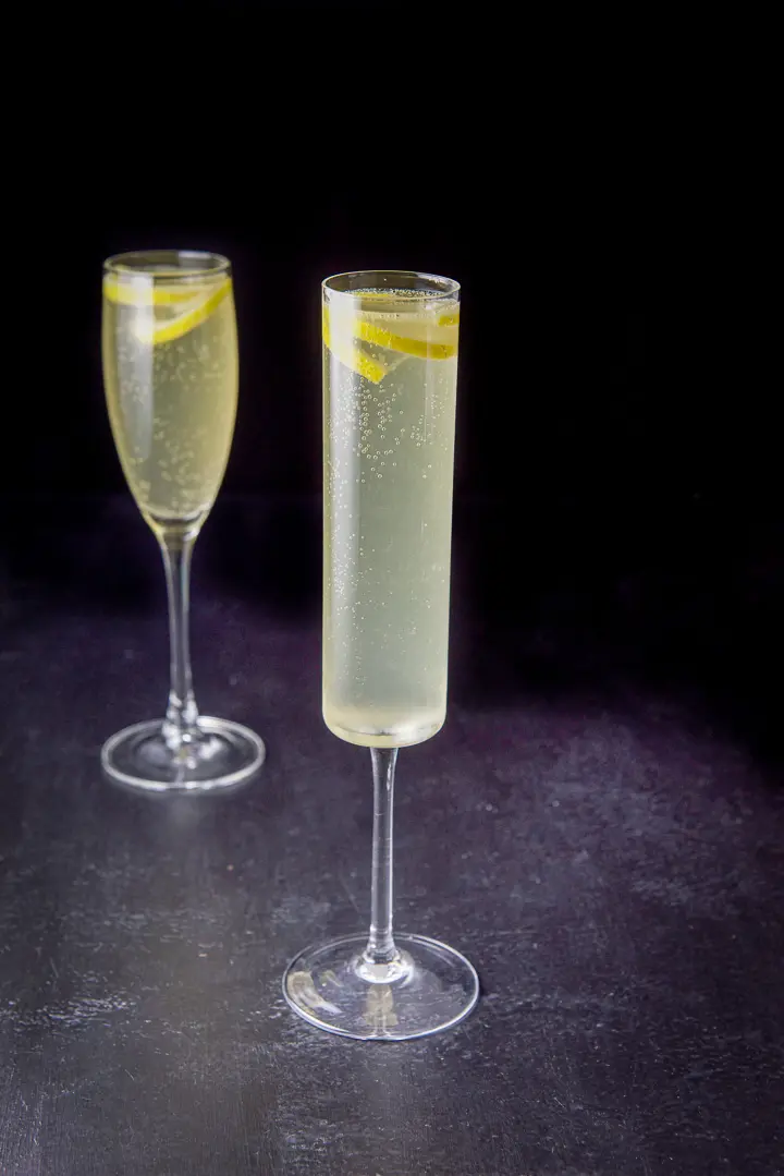 The thin champagne glass filled with the drink, the classic glass is behind it and both have lemon twists in the glasses