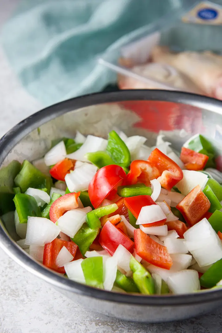 Cut up onions and peppers in a metal bowl with chicken in the background