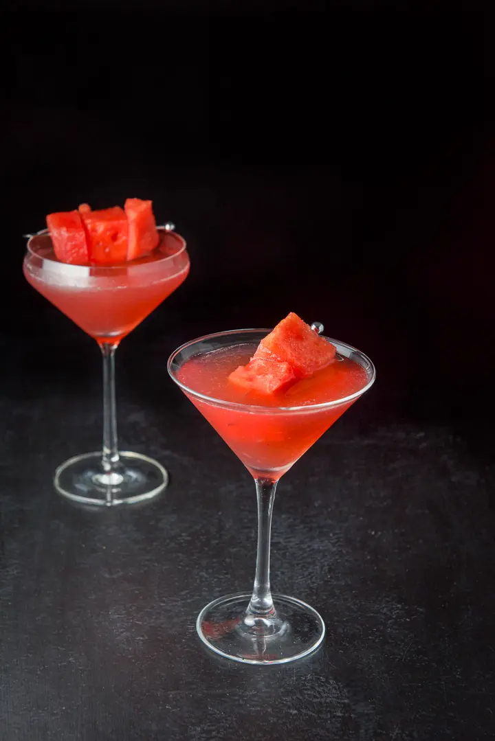 The classic martini glass in front of the shaped one filled with the cosmo with some chunks of watermelon in it