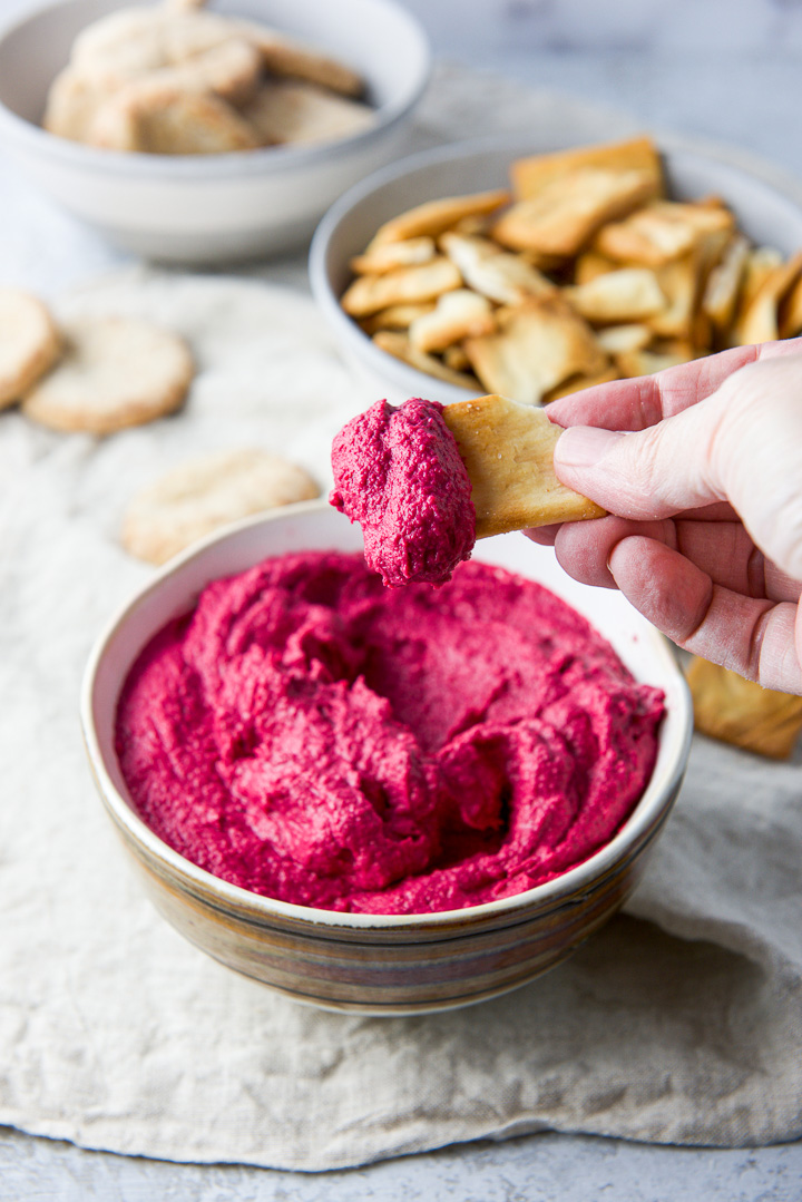 A hand holding a pita chip that was dipped in hummus over a bowl of it with chips in the background