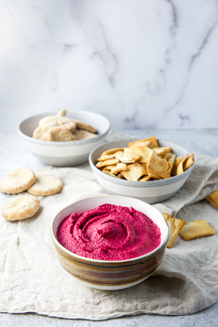 A brown and white bowl filled with hummus in a brown with pita chips and small pitas in the background