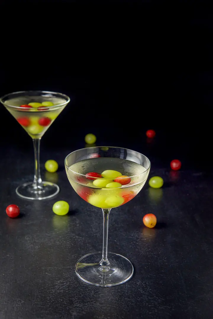 The curved glass in front of the classic martini glass. Both are filled with the cosmo with grapes in the drinks and on the table