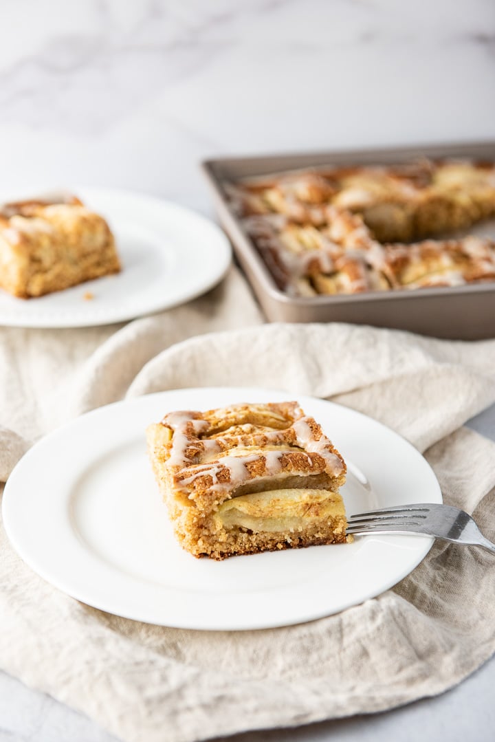A slice of cider cake on a white plate with another piece and the pan behind it