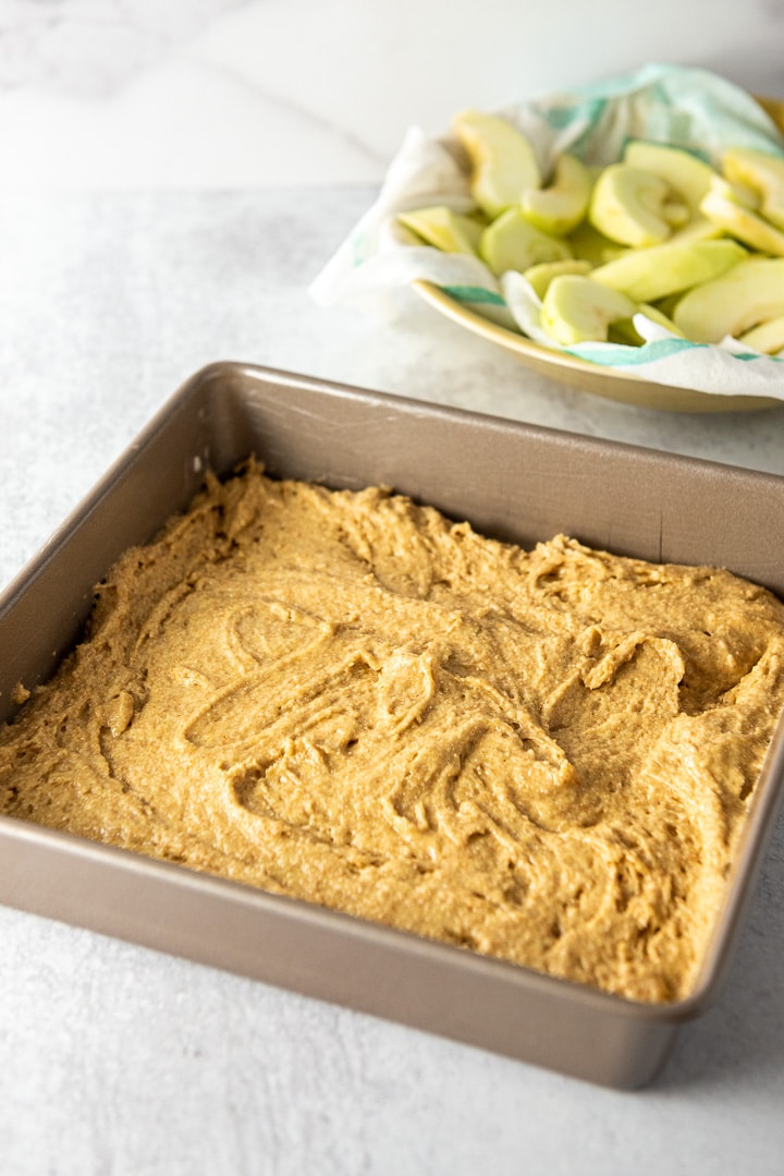Batter in a square pan with sliced apples in the background