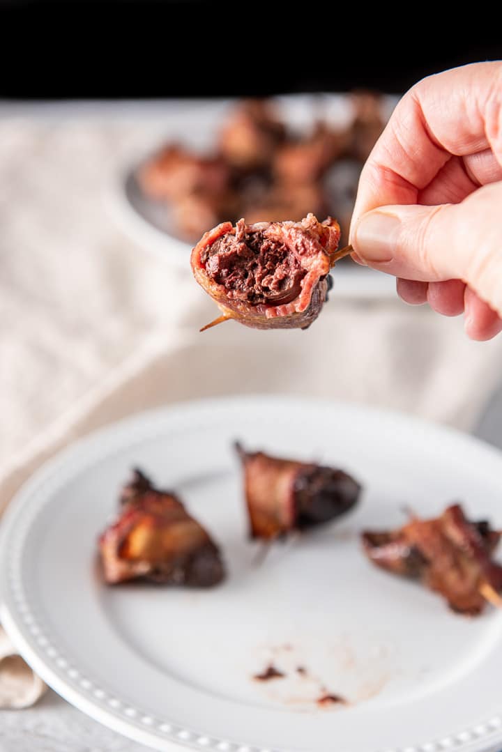 A hand holding a bacon chicken liver with a bite take out of it. There is a plate under the hand and even more livers in the background