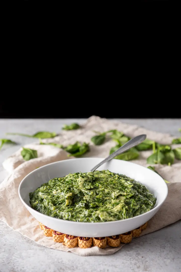 A big white bowl of creamed spinach with fresh spinach in the background