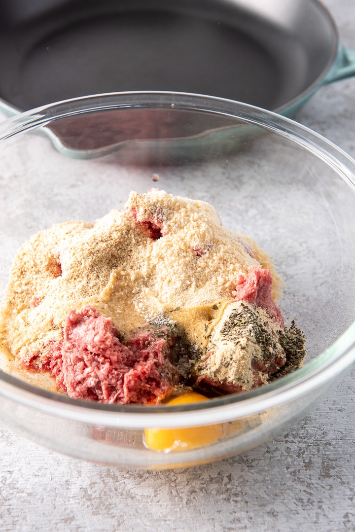 Ground beef and all the ingredients in a glass bowl with the pan in the background