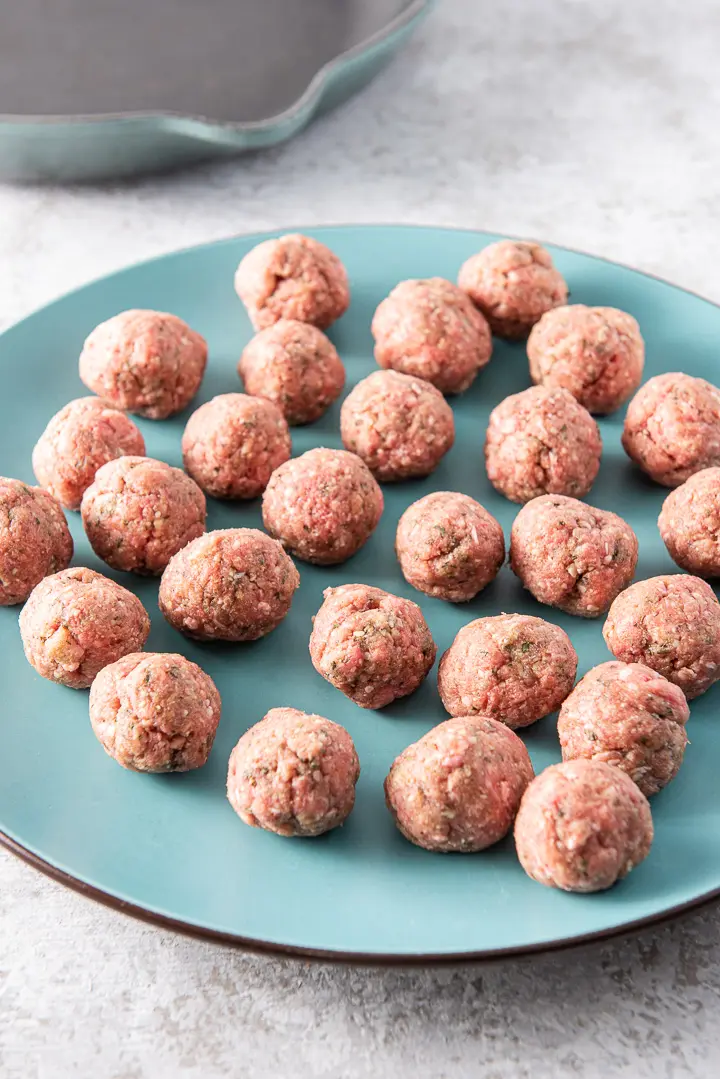 Raw meatballs on a blue platter with a cast iron pan in the background