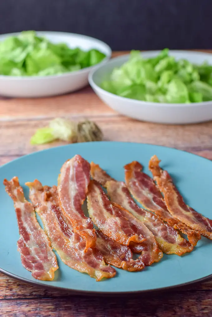 Bacon cooked and laid out on a blue plate with Boston lettuce in two white bowls in the background