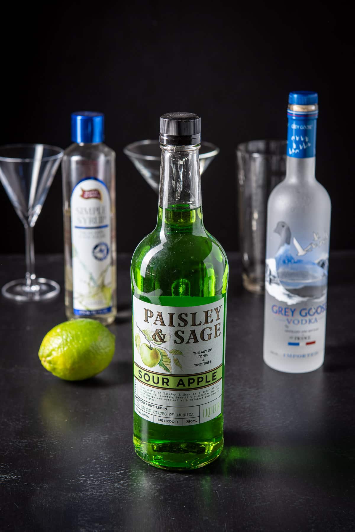 Sour apple liqueur, vodka, lime and simple syrup with the shaker and glasses in the background