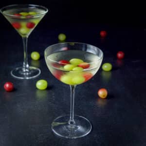 Bowl glass in front of the classic martini glass filled with the cocktail with grapes in the drink and on the table