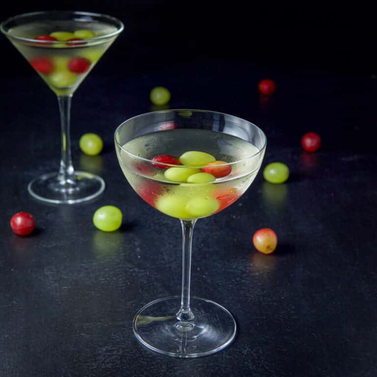 Bowl glass in front of the classic martini glass filled with the cocktail with grapes in the drink and on the table