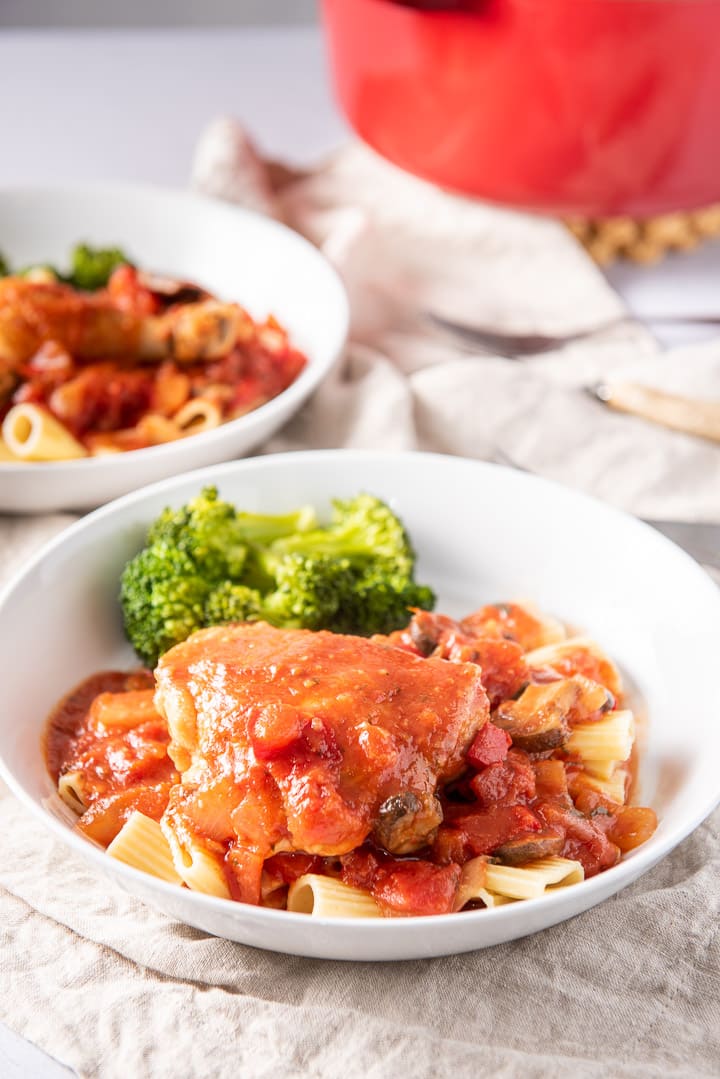 A chicken thigh on some pasta in a white plate, with another plate and pan in the background