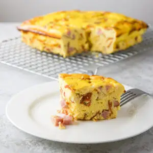 A white plate with a piece of eggy pizza gana with ham falling out of it and in the background there is the rest of it on a wire rack - square