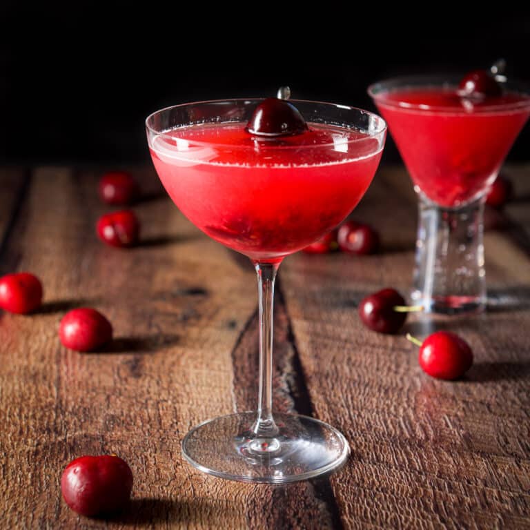 Bowl glass with the cherry cosmo in it and the bubble glass behind it. There are cherry garnishes in the glass and cherries strewn on the table