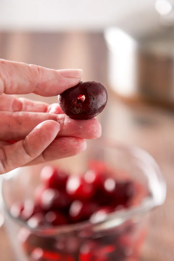 A female hand holding a pitted cherry with a hole in it over a jar of cherries
