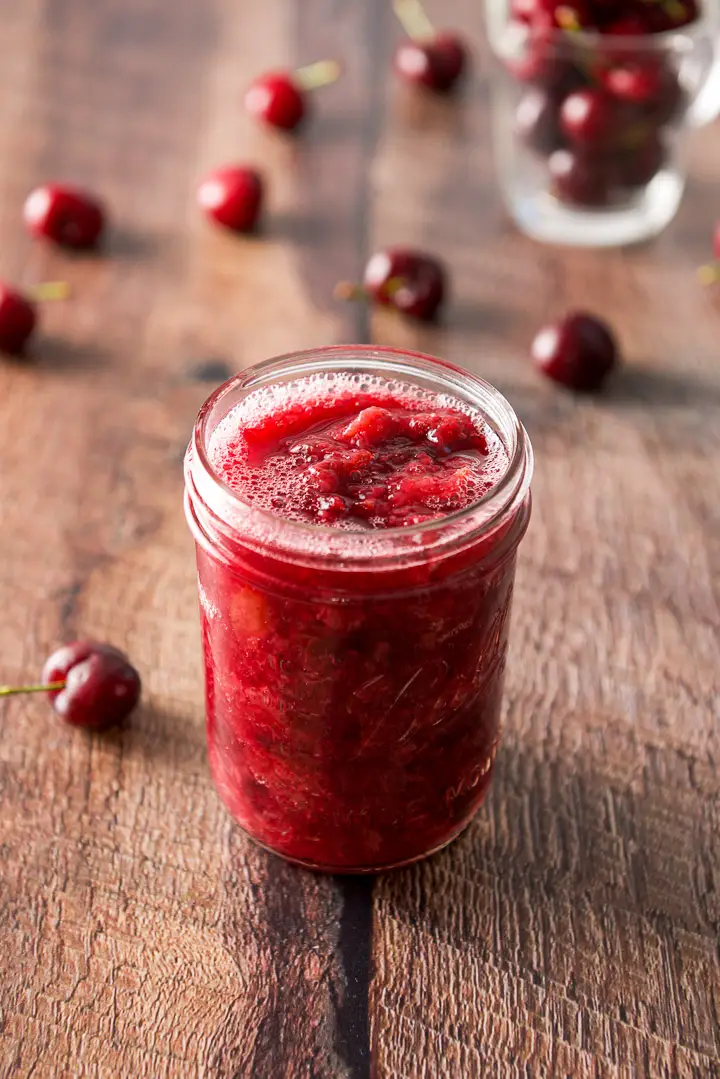 A jar of cherry sauce on the table with cherries strewn across it