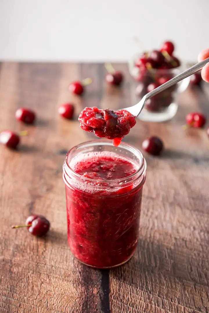A spoonful of sauce held over a jar filled with it along with cherries in a glass and on the table