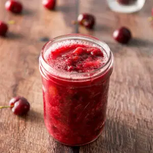 Jar of cherry sauce with cherries strewn on the table - square