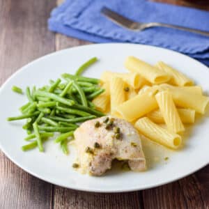A boneless skinless piece of chicken on a plate with piccata sauce, capers, ziti and green beans