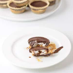 A chocolate marshmallow cup bitten into on a white plate. There are more chocolate cups behind it on a plate