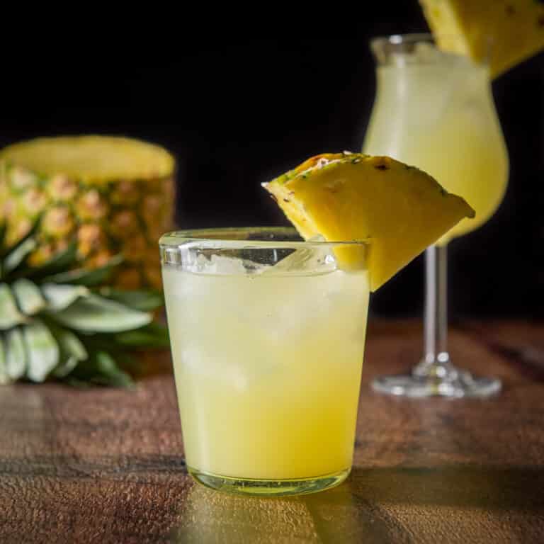 Double old fashioned glass filled with the matador with two pineapples on the side of the glass. There is a tulip glass with the cocktail and a pineapple in the background - square