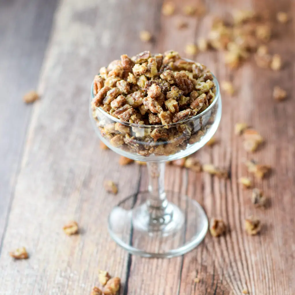 A coupe glass with candied pecans in it and a lot of pecans on the wooden table