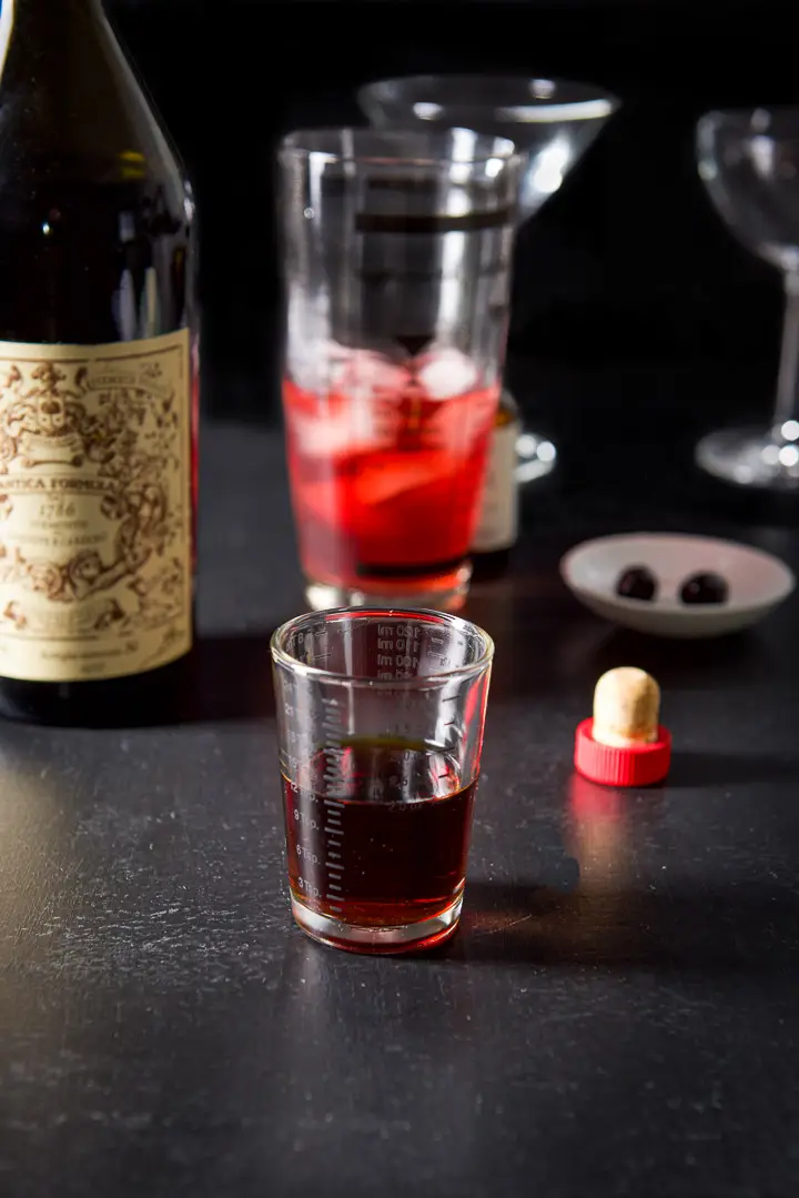 Sweet vermouth poured out with the bottle, half filled shaker, glasses and cherries in the background