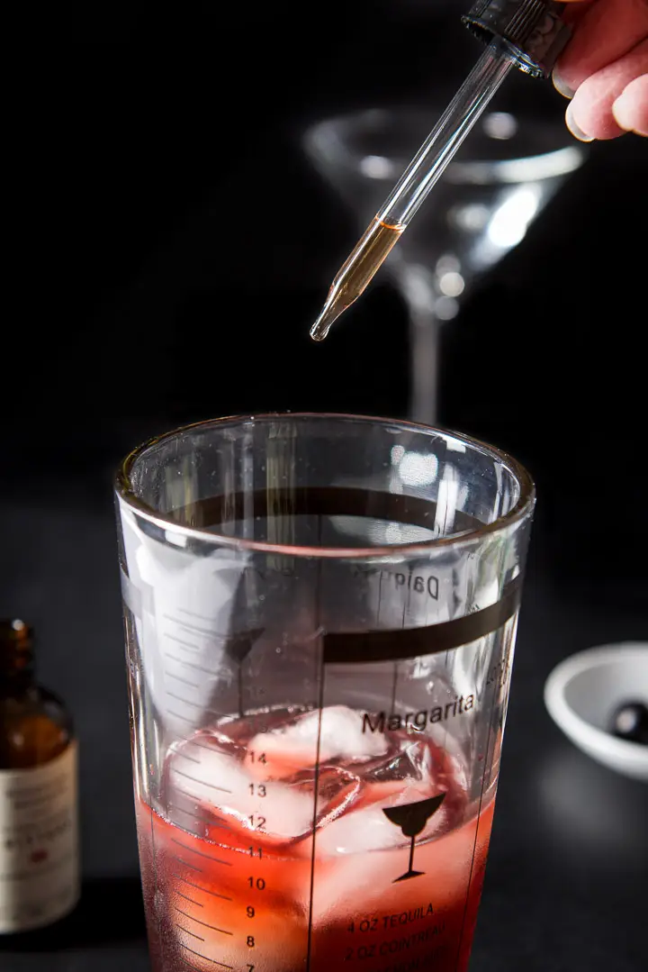 A hand holding a dropper filled halfway with cherry bitters over the cocktail shaker