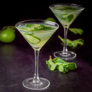 Two cucumber martinis in classic glasses with limes and mint in the background