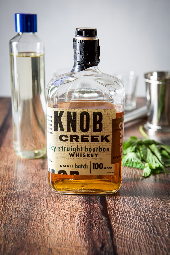 Bourbon, simple syrup and mint along with a measuring glass, muddler, glass and stainless steel glass on the table