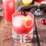 A bubble glass filled with a reddish cocktail with a lemon twist and cherries in as garnish - square
