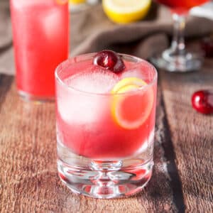 A bubble glass filled with a reddish cocktail with a lemon twist and cherries in as garnish - square