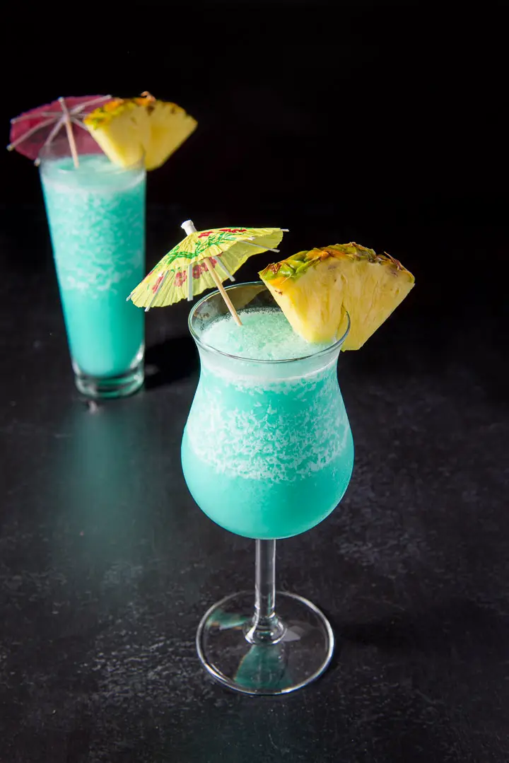 The tulip glass with the blue drink in front of the flared glass - garnished with pineapple and umbrellas