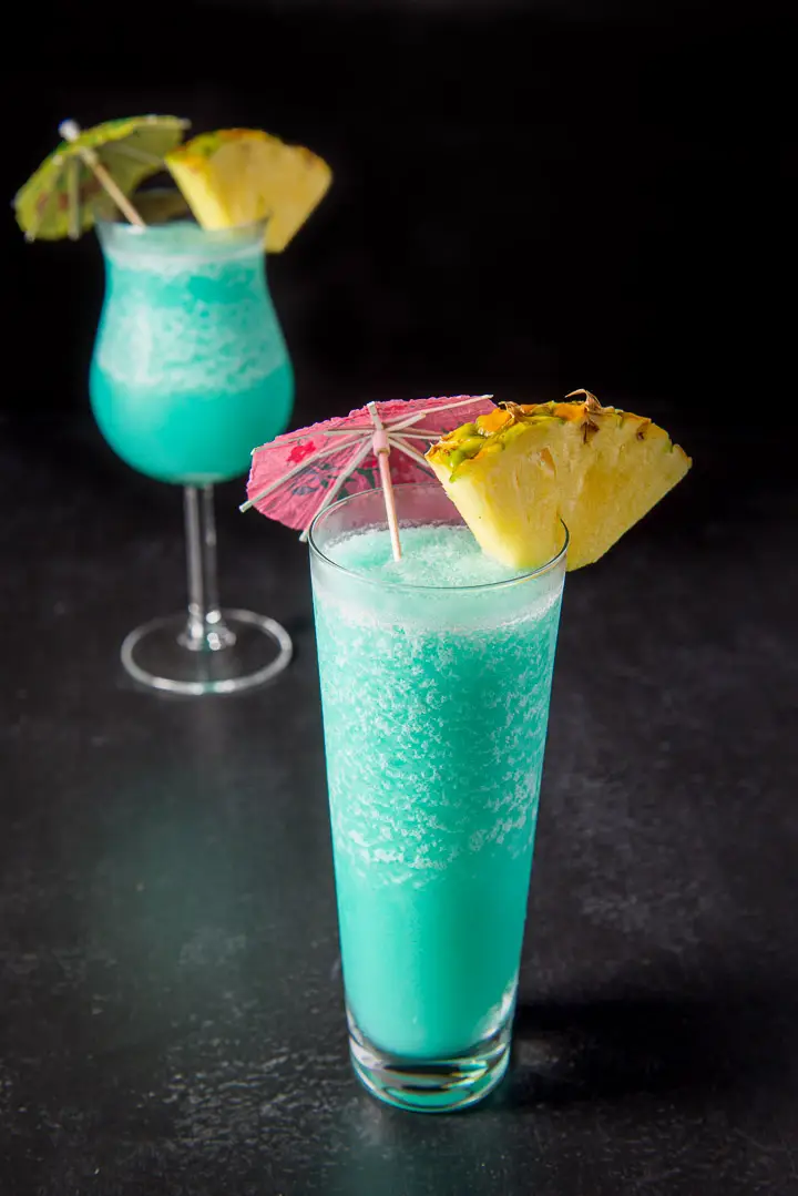 Flared glass filled with the frozen drink in front of the tulip glass. They are garnished with a pineapple wedge and umbrellas