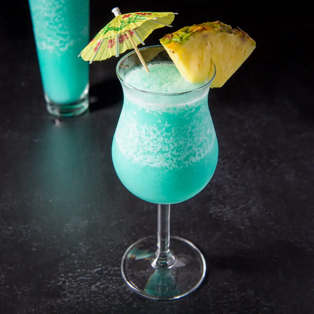 A tulip glass filled with a frozen blue drink with a pineapple wedge on the rim and a umbrella in the drink - square