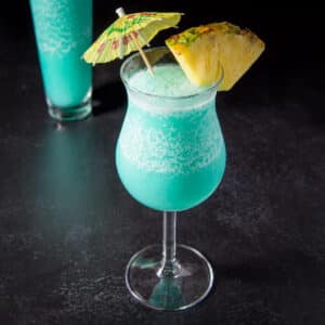 A tulip glass filled with a frozen blue drink with a pineapple wedge on the rim and a umbrella in the drink - square