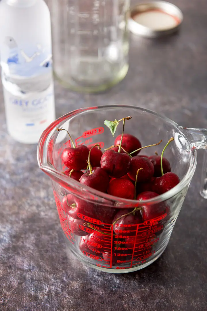 Cherries in a measuring glass with the bottle of vodka and a jar in the background